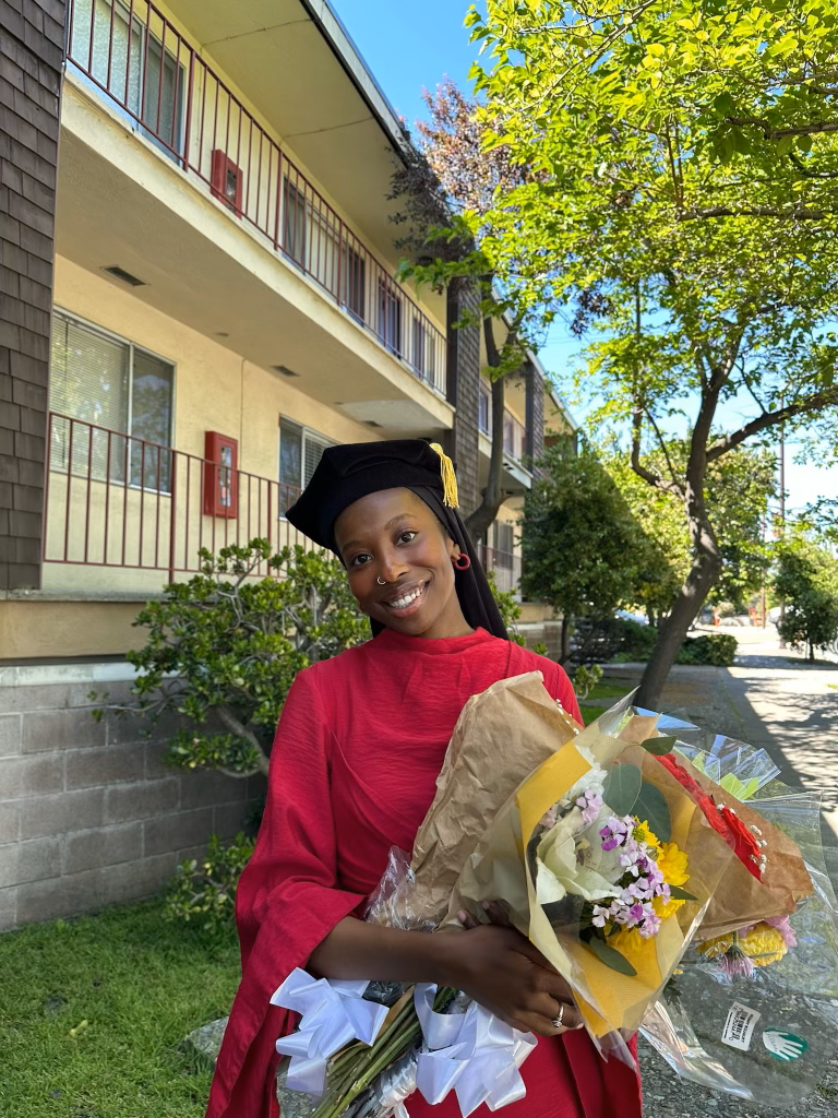 Jamilah McMillan headshot. Jamilah smiles wearing a red gown, black hijab, and graduation cap. She holds a bundle of flowers.