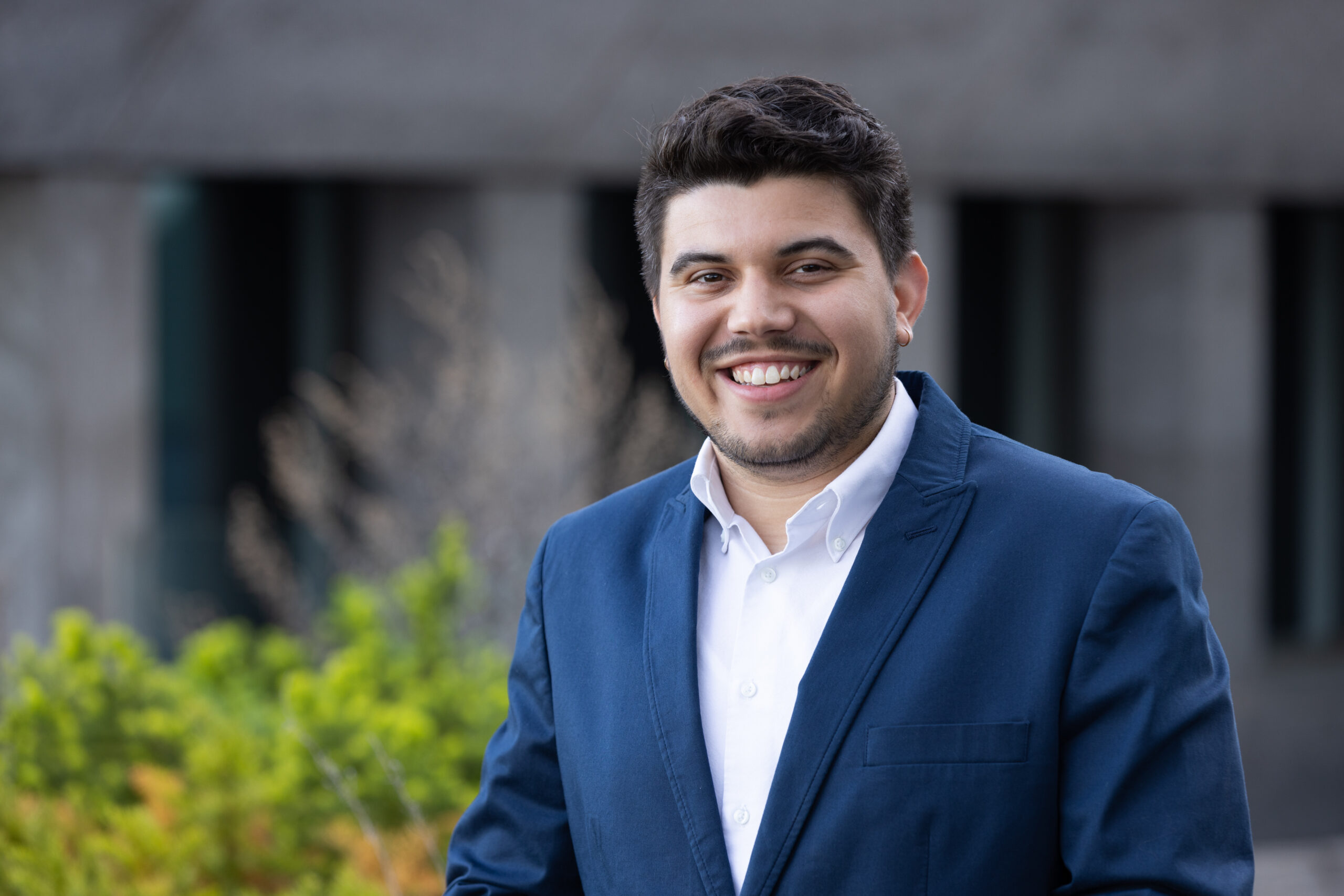 Ben Regas headshot. Ben smiles in front of a gray building and greenery. He wears a white shirt, navy blue blazer, and has short dark brown hair and facial hair.