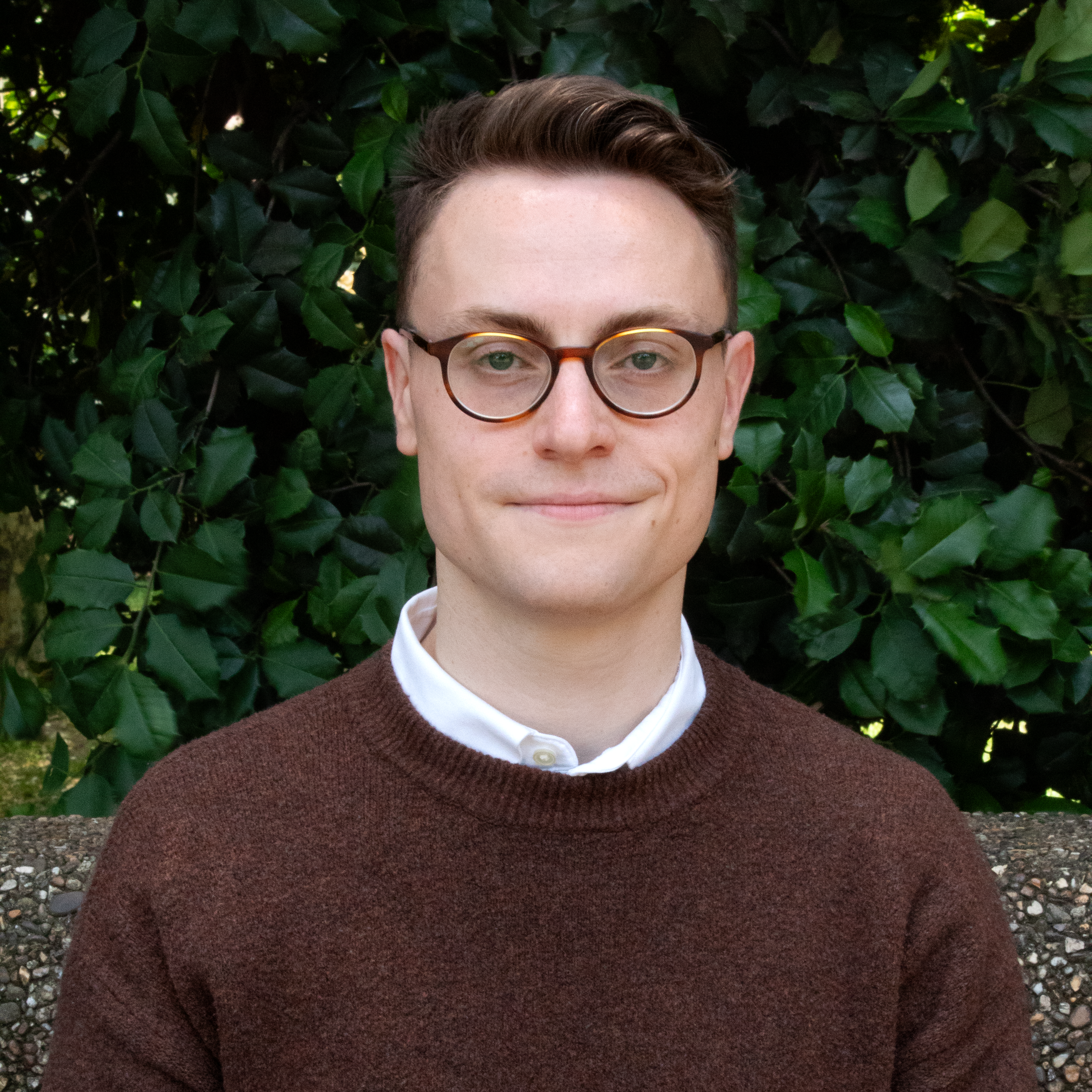 Andrew Boardman headshot. Andrew slightly smiles in front of a background of leaves. He wears a white collared shirt under a dark brown sweater and glasses. He has short brown hair.