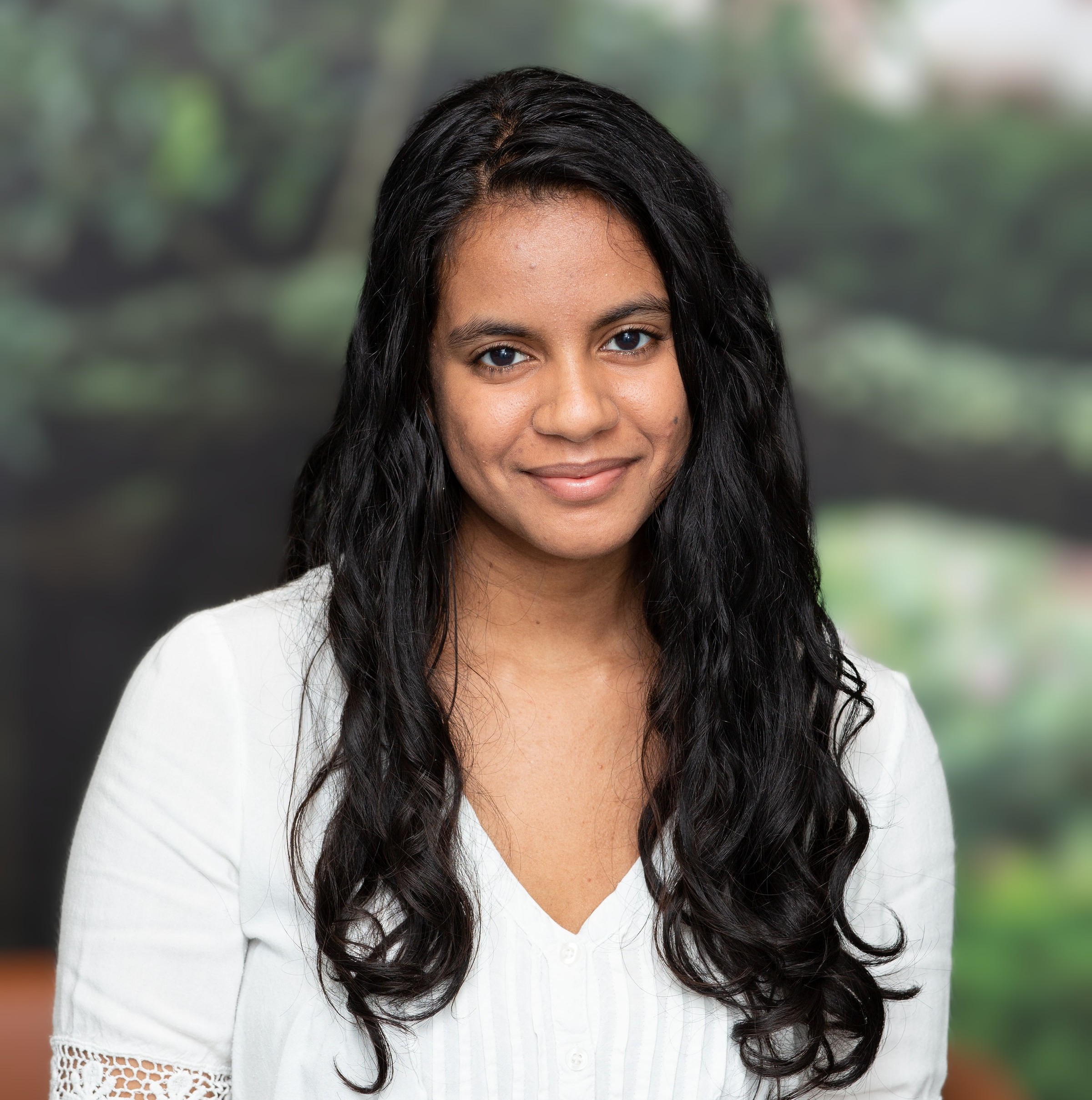 Apoorva Verghese headshot. Apoorva slightly smiles in front a blurred green background. She wears a white shirt and has long, black, wavy hair.