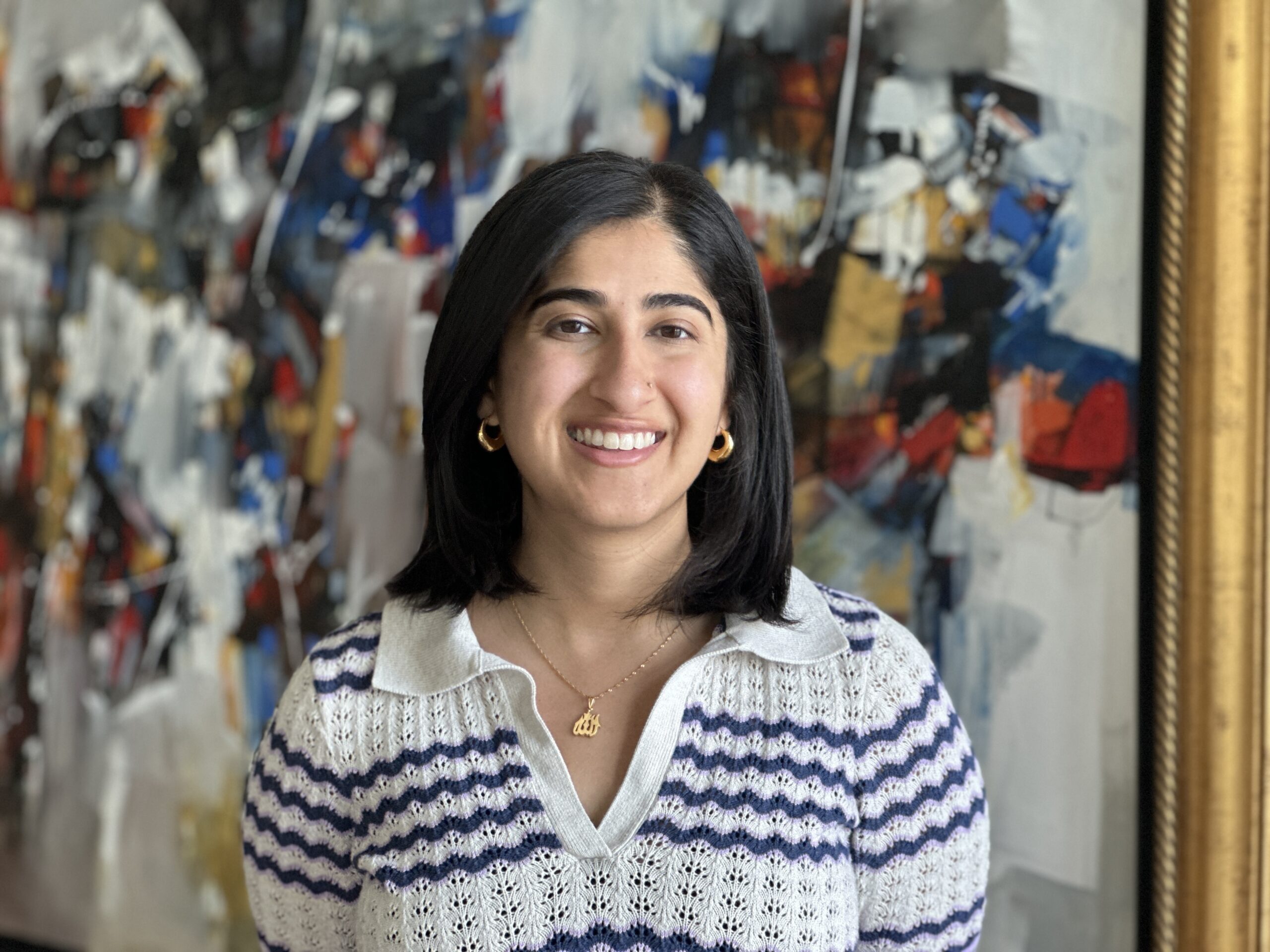 Ayeza Bajwa headshot. Ayeza smiles at the camera in front of a wall of colorful abstract art. She wears a striped blue and white sweater and has medium length straight black hair.