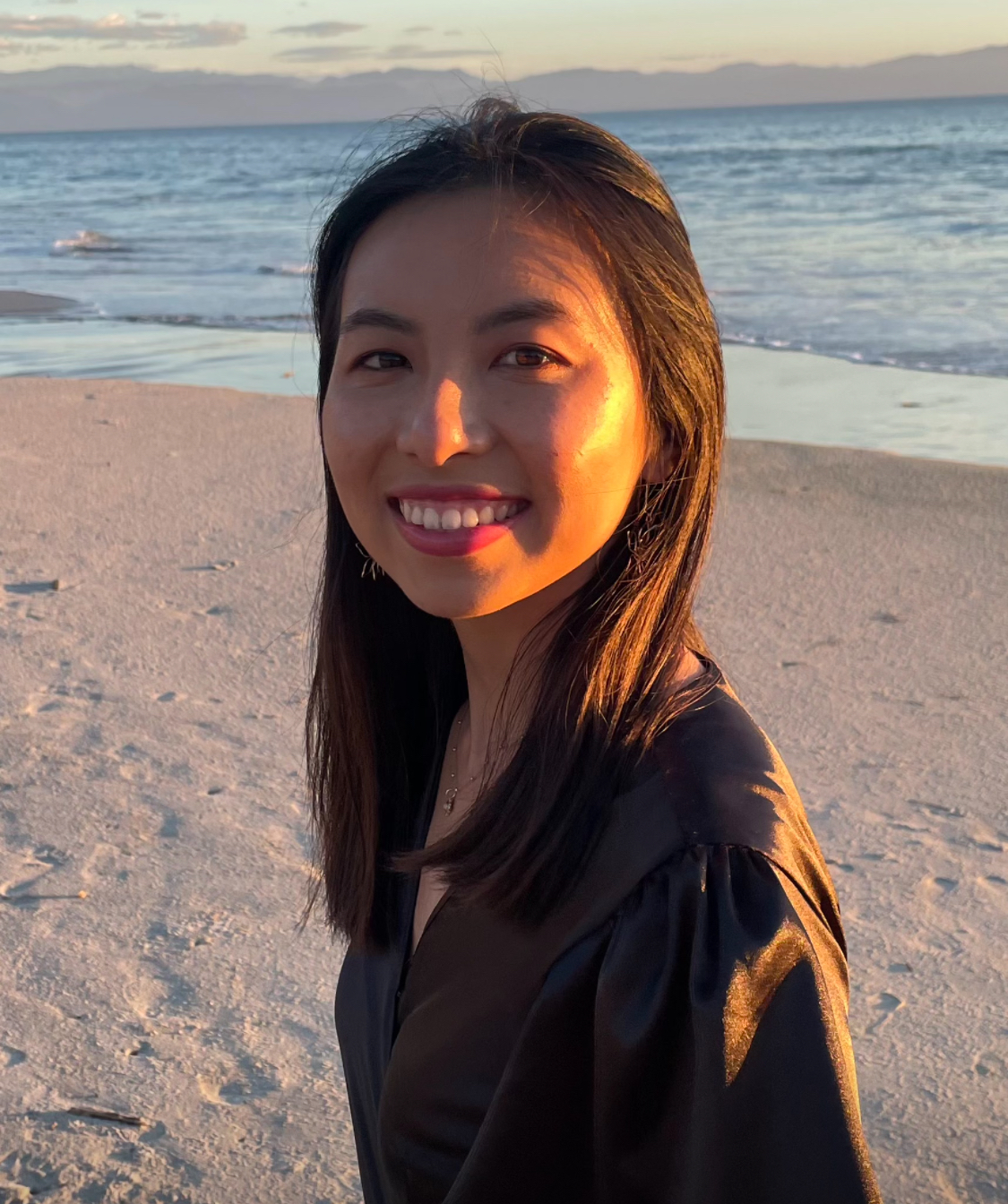 Alice Cheng headshot. Alice smiles standing on the beach in front of an incoming tide. She wears a black blouse and has long dark brown hair.