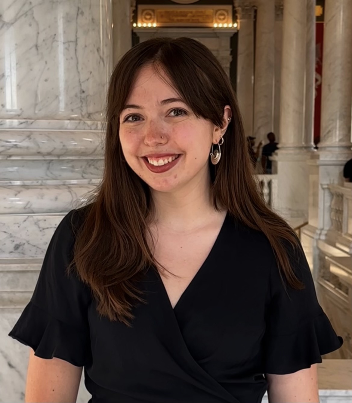 Lauren Meadows headshot. Lauren smiles standing in front of ornate marble columns. She wears a block top and has long brown hair with parted bangs.