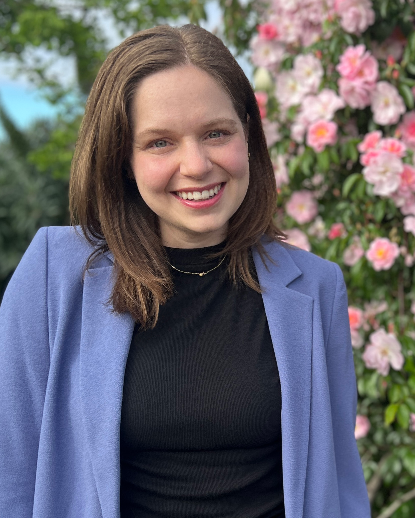 Meghan Riddlespurger headshot. Meghan smiles in front of a bloom of pink flowers. She wears a black top, blue blazer, and has medium-length brown hair.