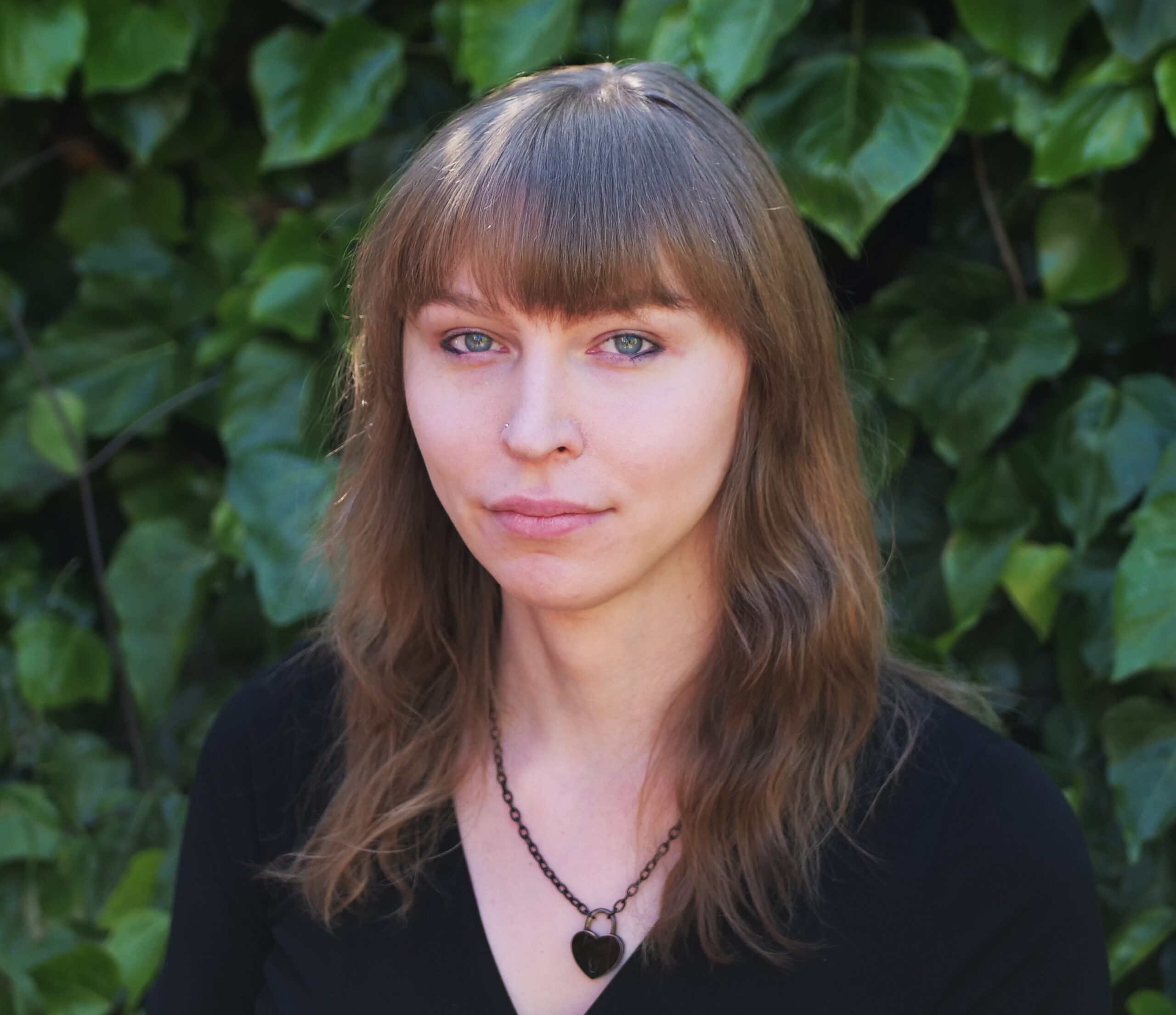 Perpetua Hilton headshot. Perpetua slightly smiles in front of a background of leaves. She wears a block top and has long light brown hair with bangs.