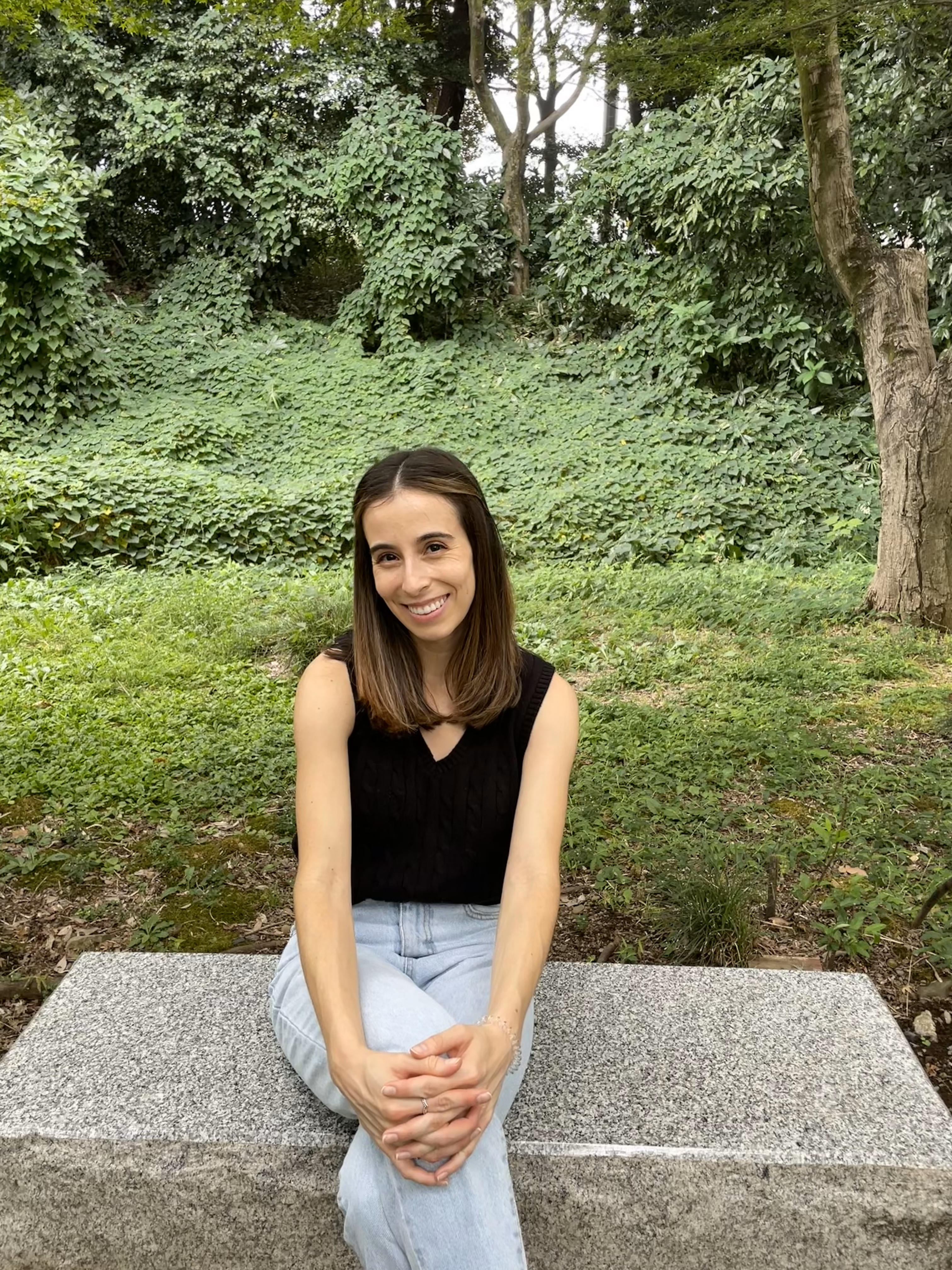 Renee Hernandez Rodriguez headshot. Renee sits on a bench and smiles in front of a background of trees and ivy. She wears a black sleeveless shirt and jeans and has long brown hair.